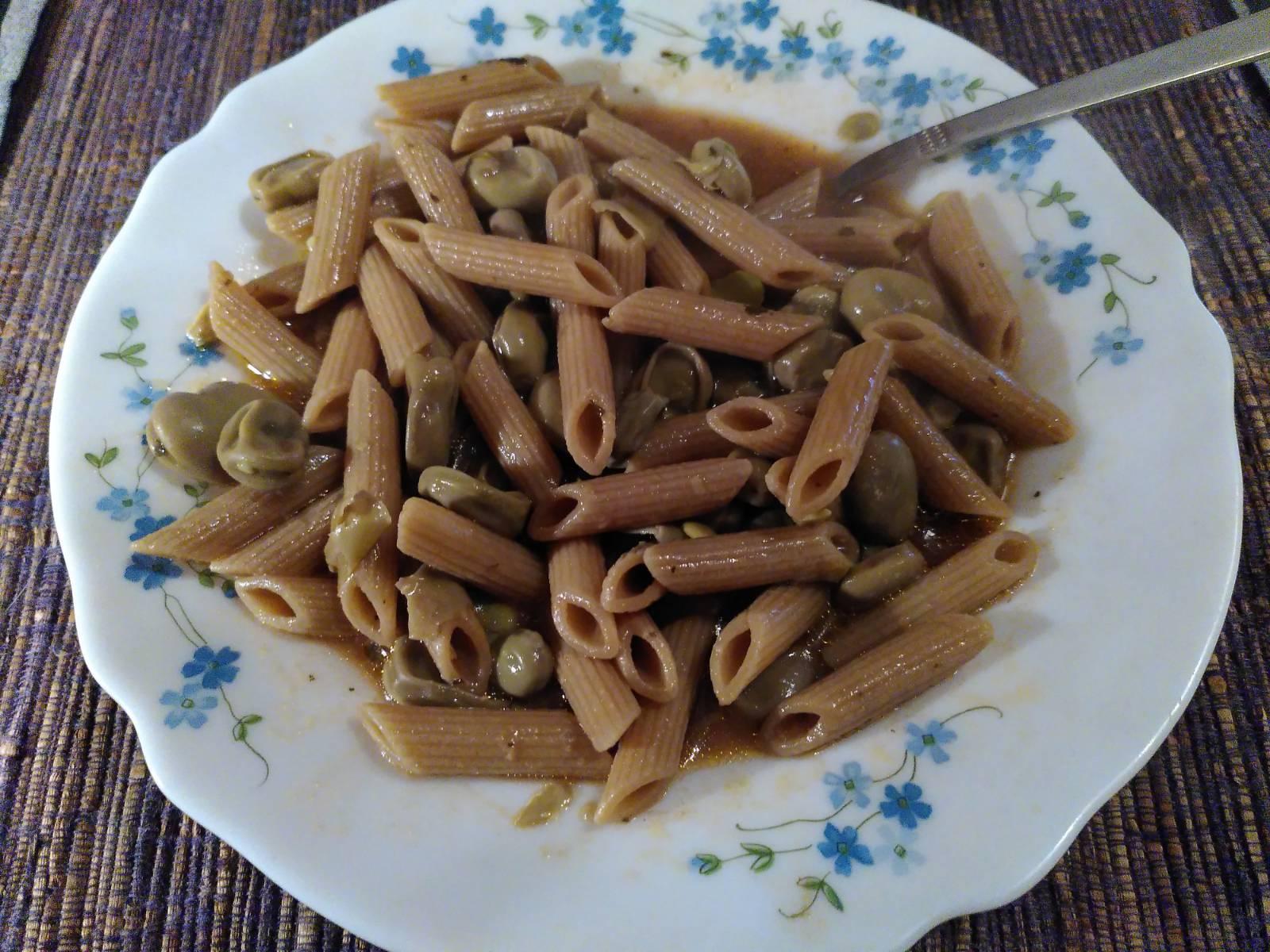 Un piatto bianco decorato con fiorellini azzurri che contiene penne integrali e fave e un liquido di colore rossastro. Sulla destra in alto, dal piatto esce un cucchiaio. Il piatto è poggiato su una tovaglietta marrone, grigia e nera