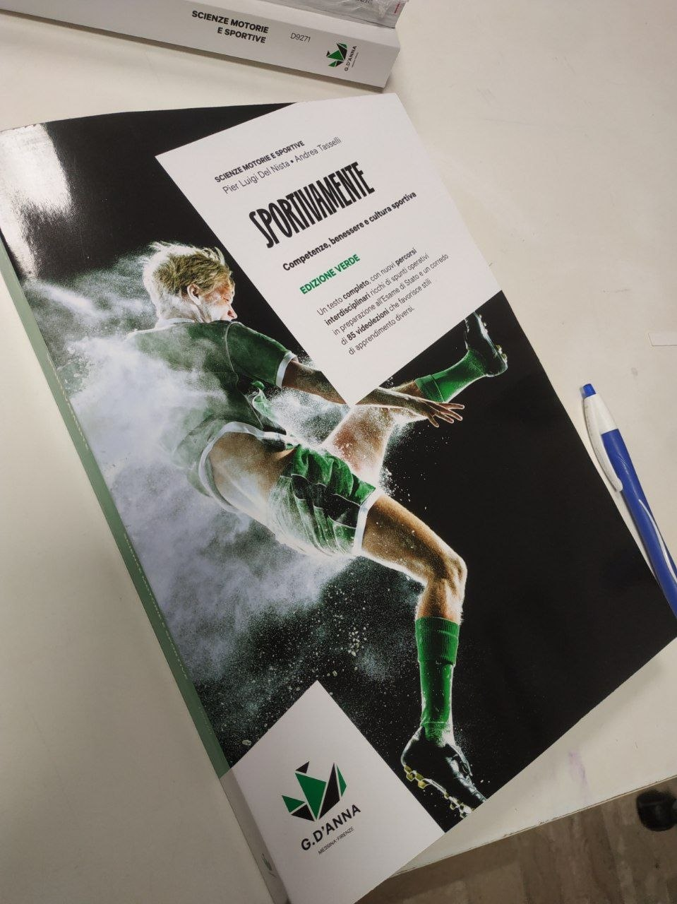 Foto di un libro scolastico sopra una scrivania bianca con una penna blu vicino.
In copertina la foto di un calciatore nell'atto di calciare in tenuta verde, la particolarità è che l'atleta è ricoperto da polvere bianca che se ne va da tutte le parti.
